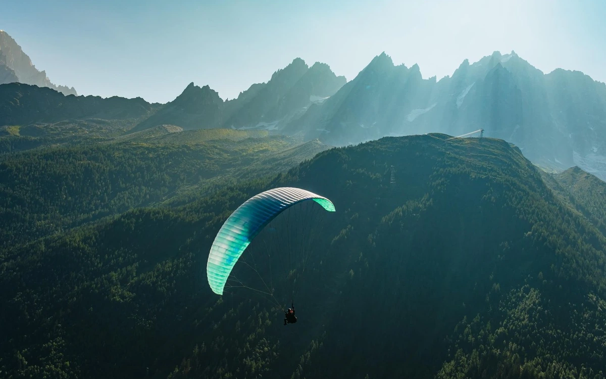 Una vela da parapendio in volo sopra creste boscose con cime illuminate dal sole sullo sfondo