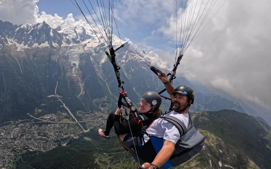 Due persone in un'imbracatura tandem in volo, con la valle e le cime bianche dietro