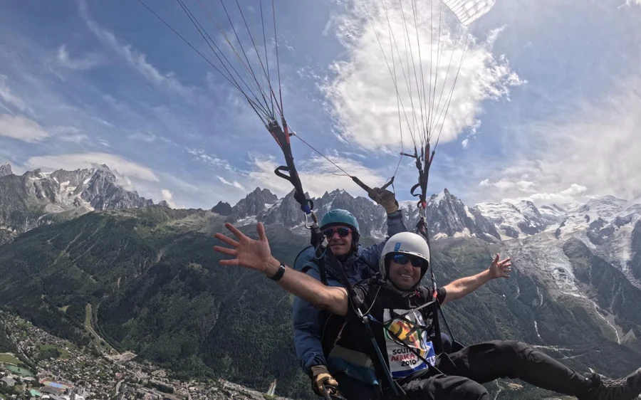 Una coppia in tandem in volo con le braccia spalancate, cime grigie e neve dietro