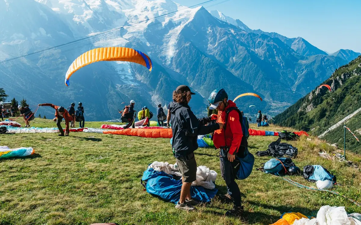 Vele da parapendio stese su un pendio erboso con piloti e passeggeri che si preparano, cime innevate sullo sfondo